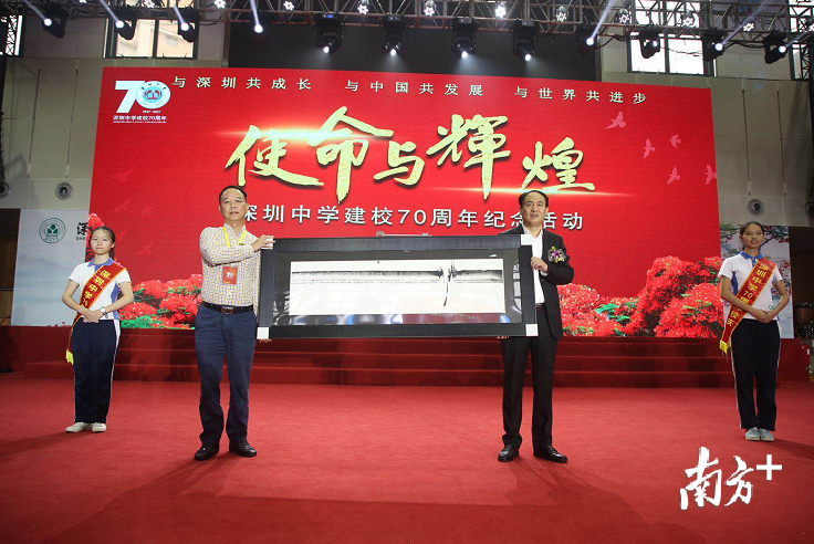 深中70华诞之际,全国各地与世界各地的校友也纷纷通过视频与微信,表达了对母校生日的祝贺与祝愿。\n在远方的祝福中,有一个特别感人的声音,它来自大洋彼岸加拿大的詹梦蕾校友。她是深圳中学77届高中毕业生,当年考取大学的三个应届毕业生之一。今年五月,她携身为联合国艺术大使的丈夫何靖恒先生回深圳探望母校,并决定把先生所创作的代表联合国理念的书画艺术作品“環宇一心”作为华诞贺礼,敬赠母校。10天前,就在他们准备返程时,詹梦蕾校友突感不适,到医院检查,已是癌症晚期。\n无情的疾病阻挡了詹梦蕾校友夫妇的脚步,但没有阻止他们的校缘深情。几天前,他们已经将这幅杰出作品快寄回国,委托同学甘平校友来完成他们的至高心意。\n深中77届校友甘平和朱华伟接受这份来自大洋彼岸的心意。