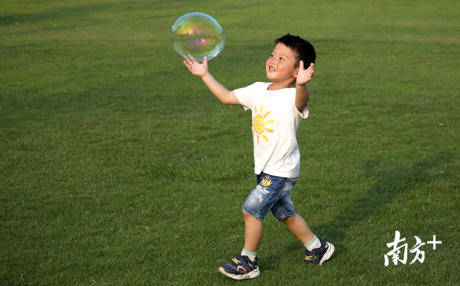金山公园草地上，小男孩一边奔跑，一边小心翼翼呵护着眼前的泡泡，脸上挂着童真的微笑。