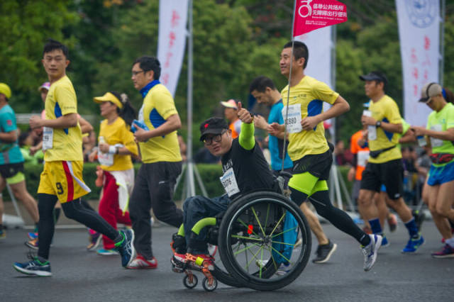 2018年3月18日，一名残疾运动员推着轮椅车参赛，举旗呼吁无障碍出现。资料图